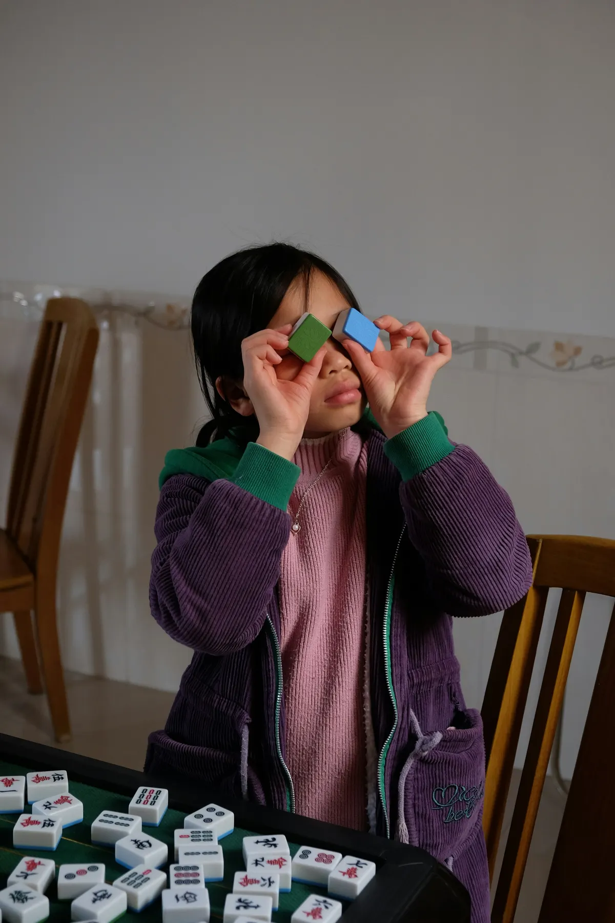 A young player playfully holding two Mahjong tiles up to her eyes while learning the game